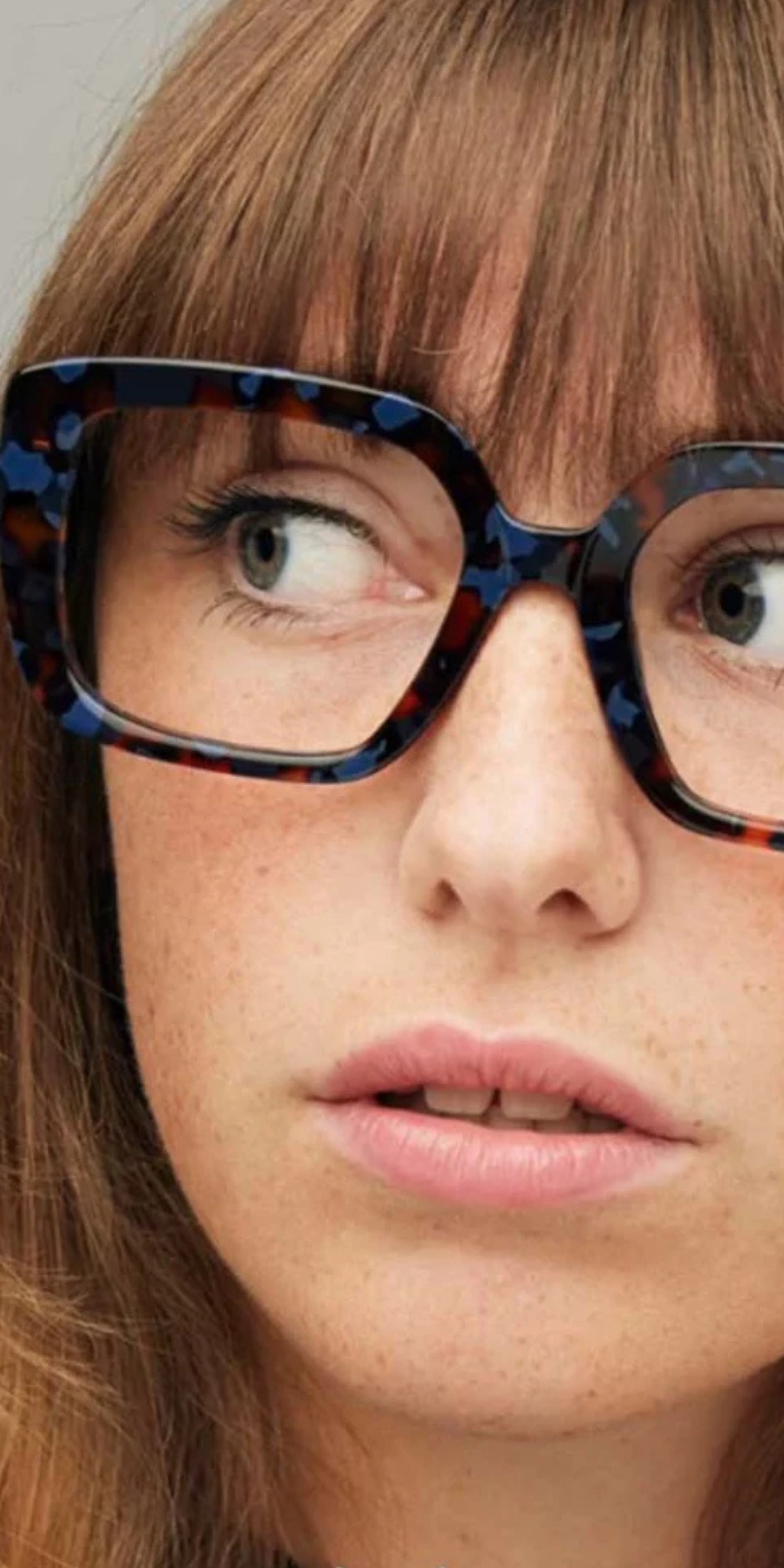 Close-up of a woman with red hair wearing large, patterned glasses, looking to the side with her mouth slightly open.