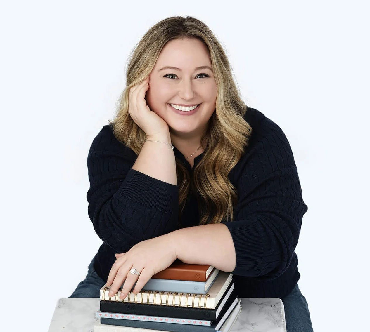 A smiling woman with long wavy blonde hair, wearing a black sweater, resting her head on her hand, sitting at a table with a stack of notebooks and journals, against a plain light background.