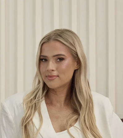 A smiling young woman with blonde hair wearing a white blouse, sitting on a beige chair in a neutral room.