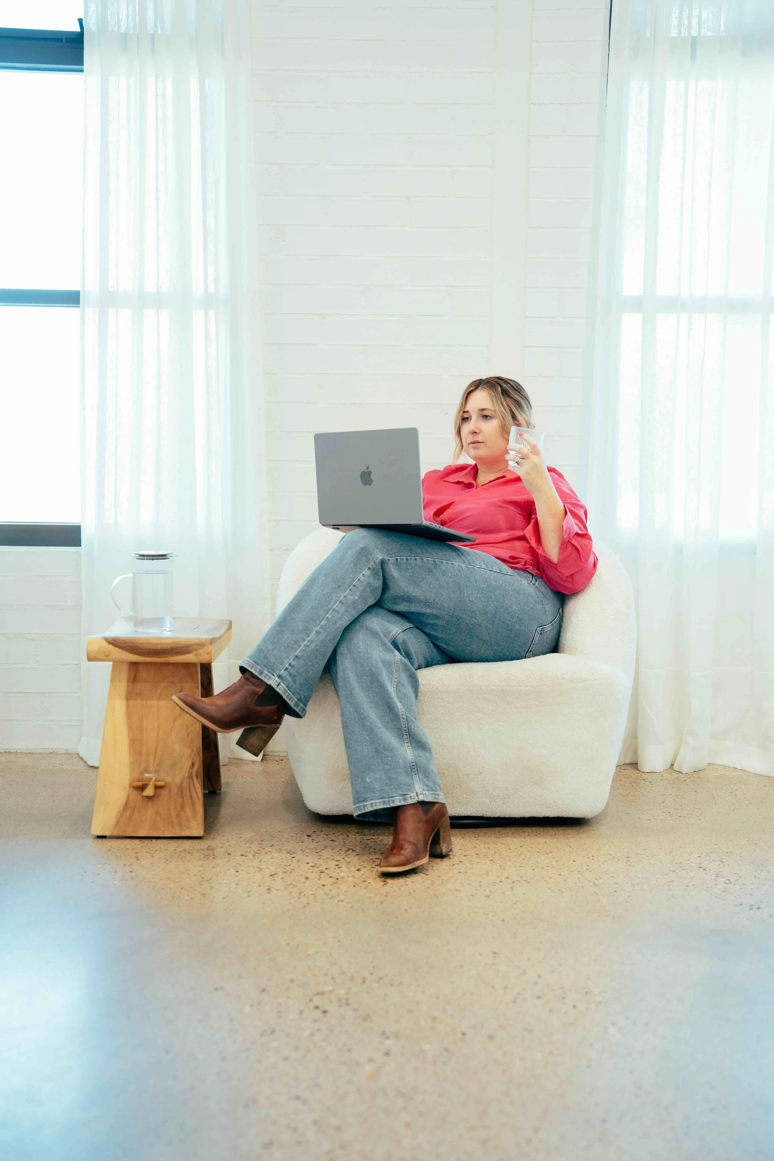 A woman sitting on a white armchair in a bright room, holding a glass of water and working on a laptop placed on her lap.
