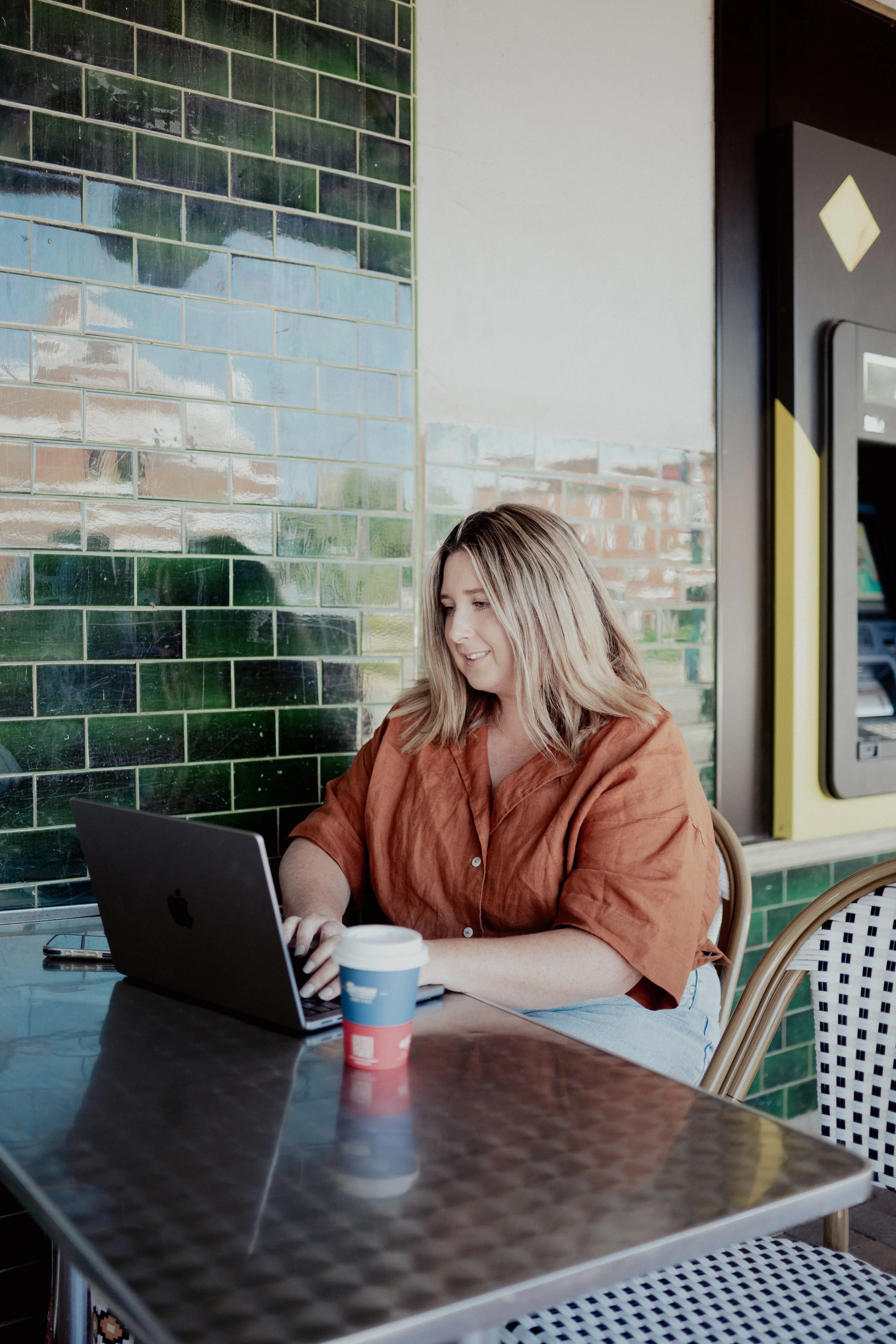 A woman with blonde hair wearing an orange shirt is sitting at a table, working on a laptop, with a coffee cup nearby, against a background of a tiled green wall and a vending machine.