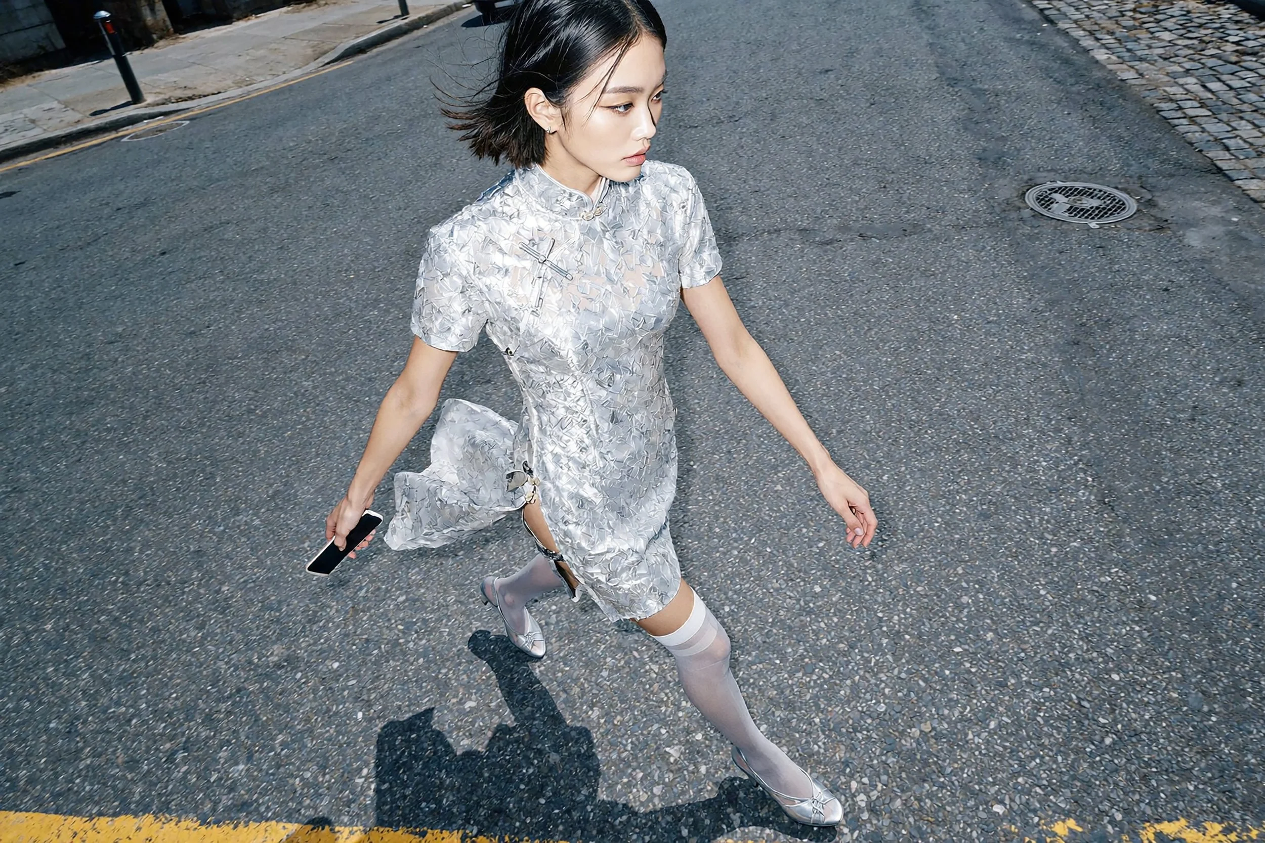 Model Jun Xeda in an editorial street photoshoot, dressed in a new-age white silver Chinese Qipao dress with matching white stockings. Dynamic angle shot that focuses on her movement as she crosses an urban concrete street. Produced by digital fashio