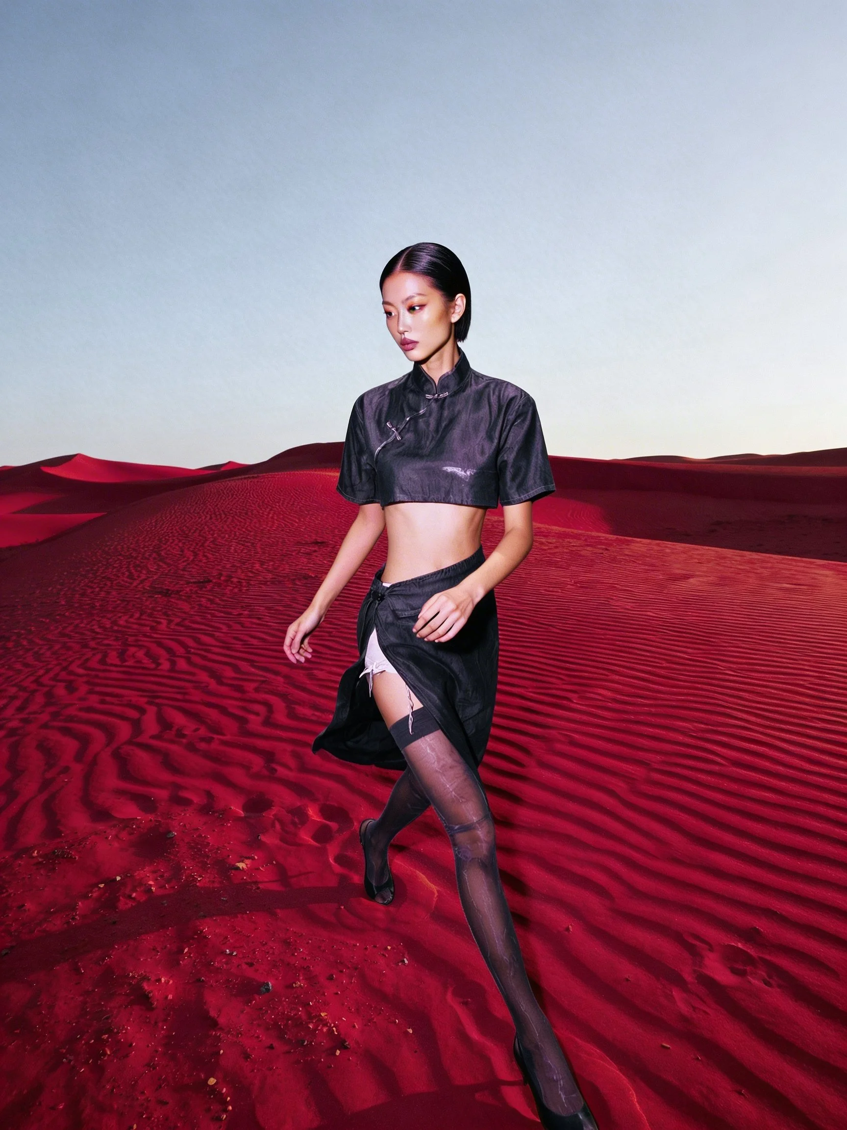 Model Xia Iseri posing in a new-age Chinese Qipao in a deep-red sandy desert like environment. The outfit consists of a cropped top, matching skirt and black stockings, complete with oriental details that fuses modernity with tradition.