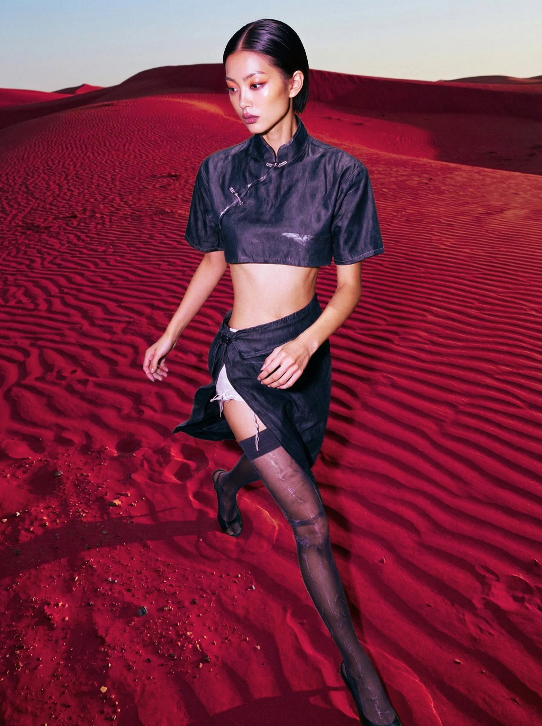Model Xia Iseri posing in a new-age Chinese Qipao in a deep-red sandy desert like environment. The outfit consists of a cropped top, matching skirt and black stockings, complete with oriental details that fuses modernity with tradition.