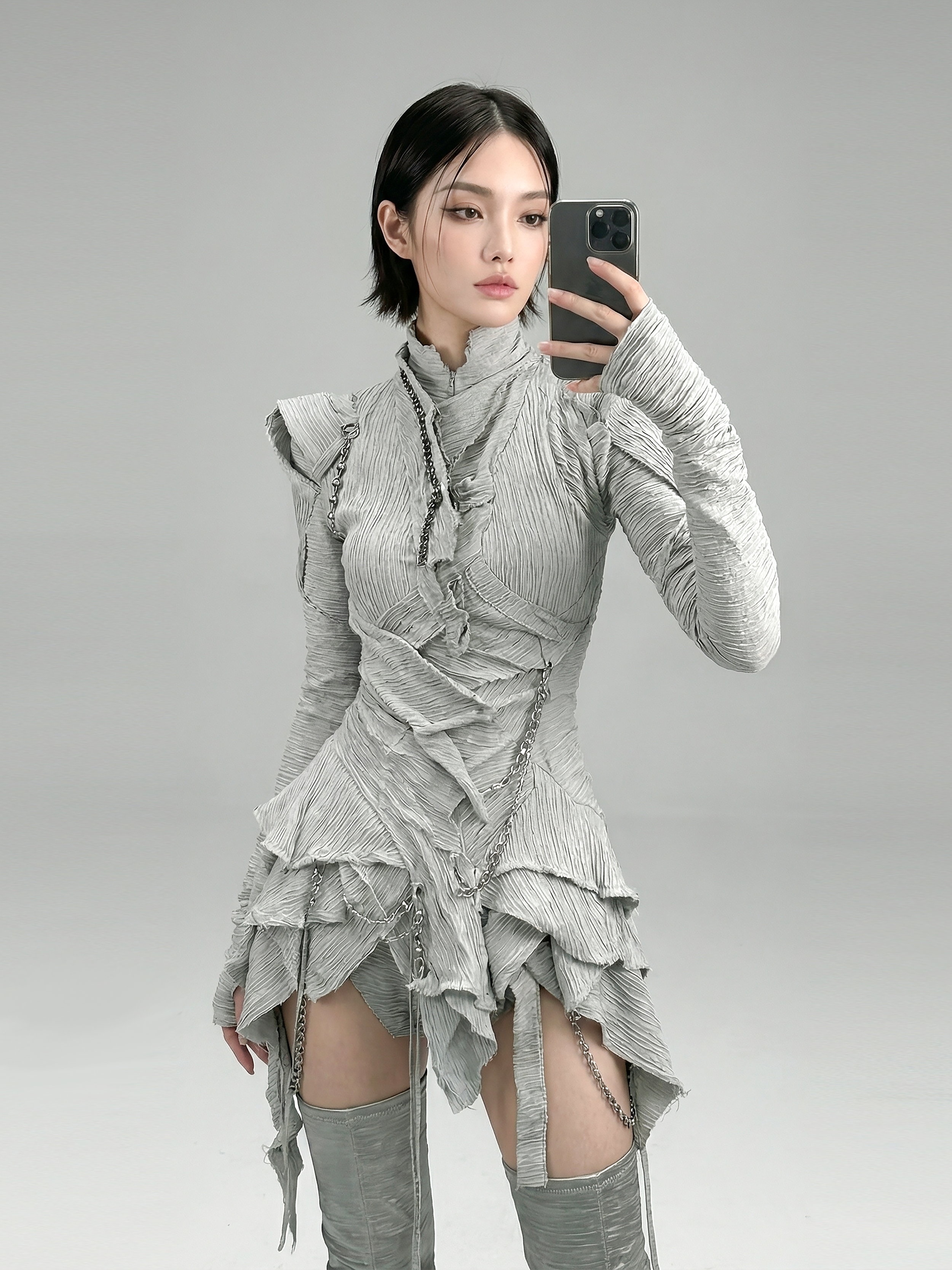 Model and brand ambassador Jun Xeda wearing an otherworldly light gray textured dress that is layered with fitted bodice, expressed ruffles and a draped hemline. She is wearing thigh high silver boots with a tabi toe design, for digital fashion brand