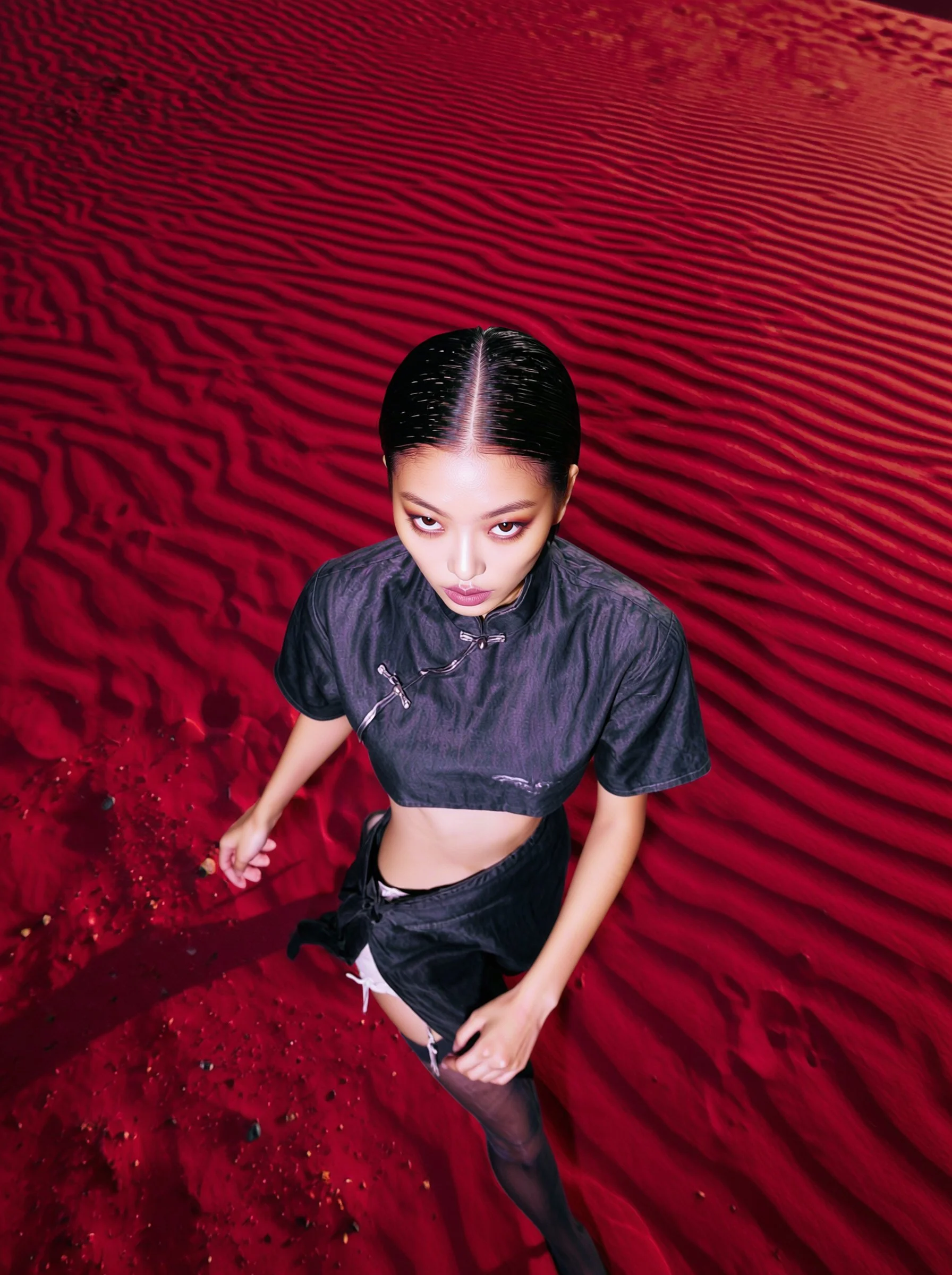 Model Xia Iseri posing in a new-age Chinese Qipao in a deep-red sandy desert like environment. The outfit consists of a cropped top, matching skirt and black stockings, complete with oriental details that fuses modernity with tradition.