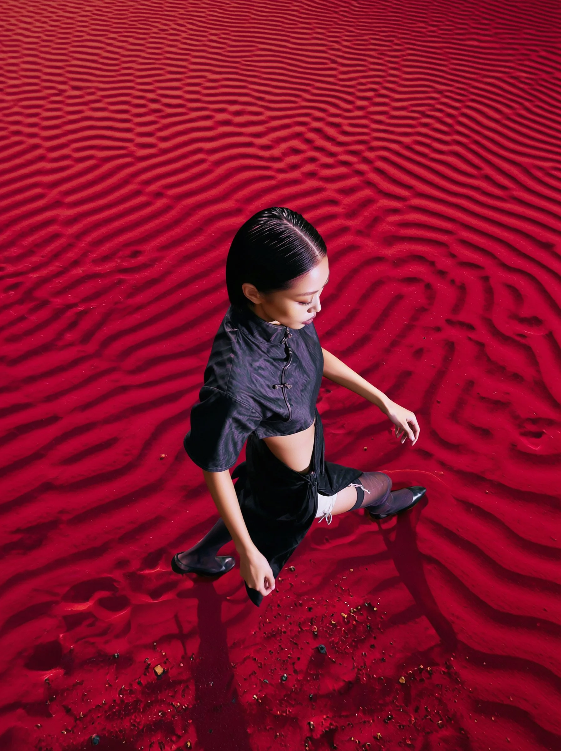 Model Xia Iseri posing in a new-age Chinese Qipao in a deep-red sandy desert like environment. The outfit consists of a cropped top, matching skirt and black stockings, complete with oriental details that fuses modernity with tradition.