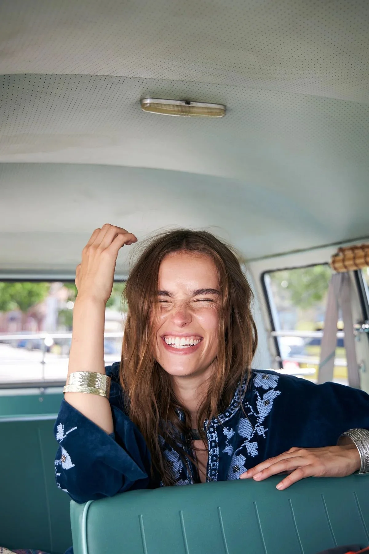 A woman with long brown hair and a wide smile, sitting inside a vintage van. She is wearing a dark blue embroidered top and a silver bracelet, with her eyes closed in a laughing expression.