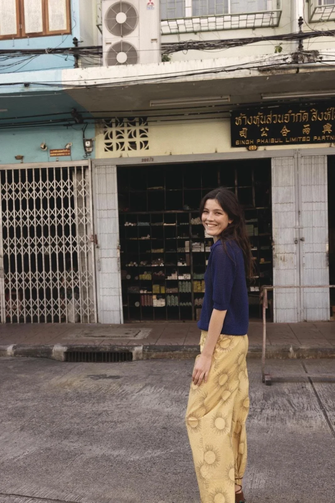 A woman with long dark hair is standing on a street, smiling and looking at the camera. She is wearing a blue top and yellow pants with a circular pattern. Behind her, there are storefronts with metal grates and a black sign with yellow text in Thai and English.