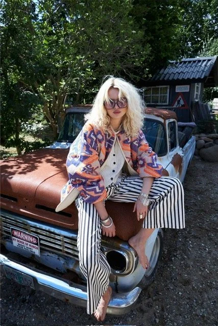 A woman with curly blonde hair, wearing sunglasses, a patterned jacket, a white top, and striped pants, sitting on the hood of a vintage rusty pickup truck outdoors.