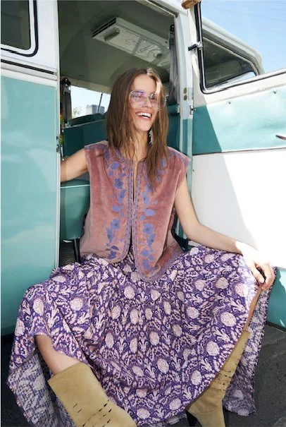 A woman sitting at the door of a vintage teal and white camper van, smiling, wearing glasses, a patterned purple skirt, a sleeveless vest, and tan boots.