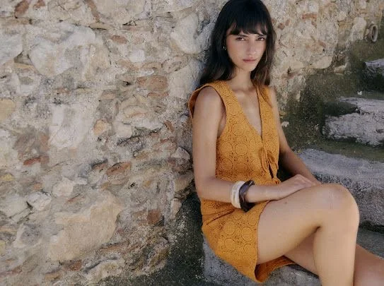 A young woman sitting on stone steps beside a stone wall, wearing an orange dress and bracelets.
