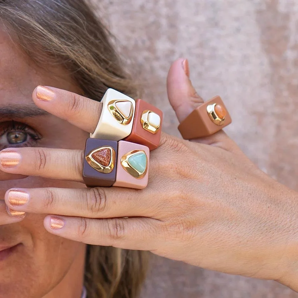 A woman's fingers and face are partially visible, showcasing five large, colorful rings on her fingers. The rings are chunky with geometric designs, featuring various colors and gold accents.