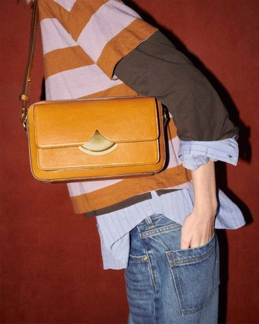 Person wearing a striped sweater, layered over a button-up shirt, with jeans, holding a mustard yellow clutch bag against a red background.