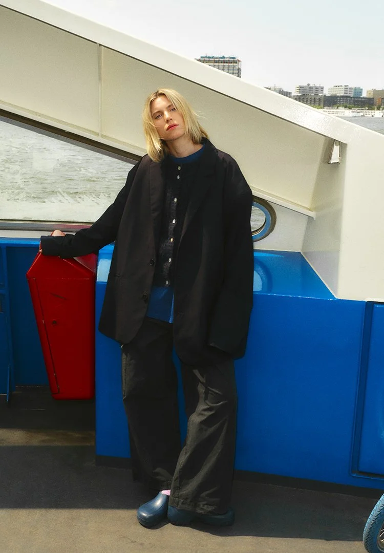 A young woman with shoulder-length blonde hair stands on a boat deck, leaning against a red and blue structure, with a city skyline in the background.