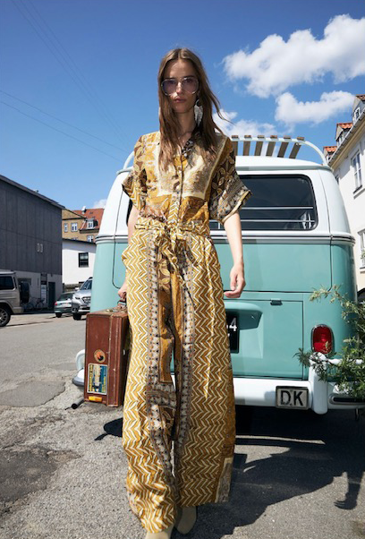 A woman in yellow patterned jumpsuit with sunglasses and a suitcase, standing in front of a vintage teal van on a sunny day.