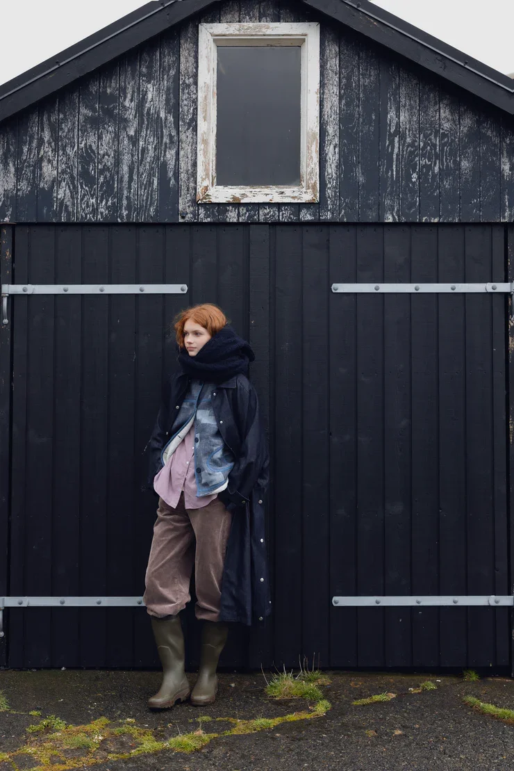 A young woman with red hair wearing a black scarf, blue jacket, pink shirt, brown pants, and green rubber boots standing in front of a black wooden barn with a small boarded-up window.