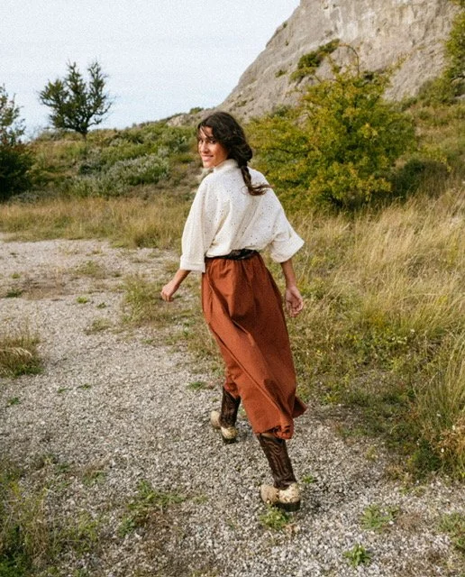 Woman walking outdoors on gravel path with grassy hills and rocky cliffs in background, wearing a white blouse, brown wide-leg pants, and patterned shoes.
