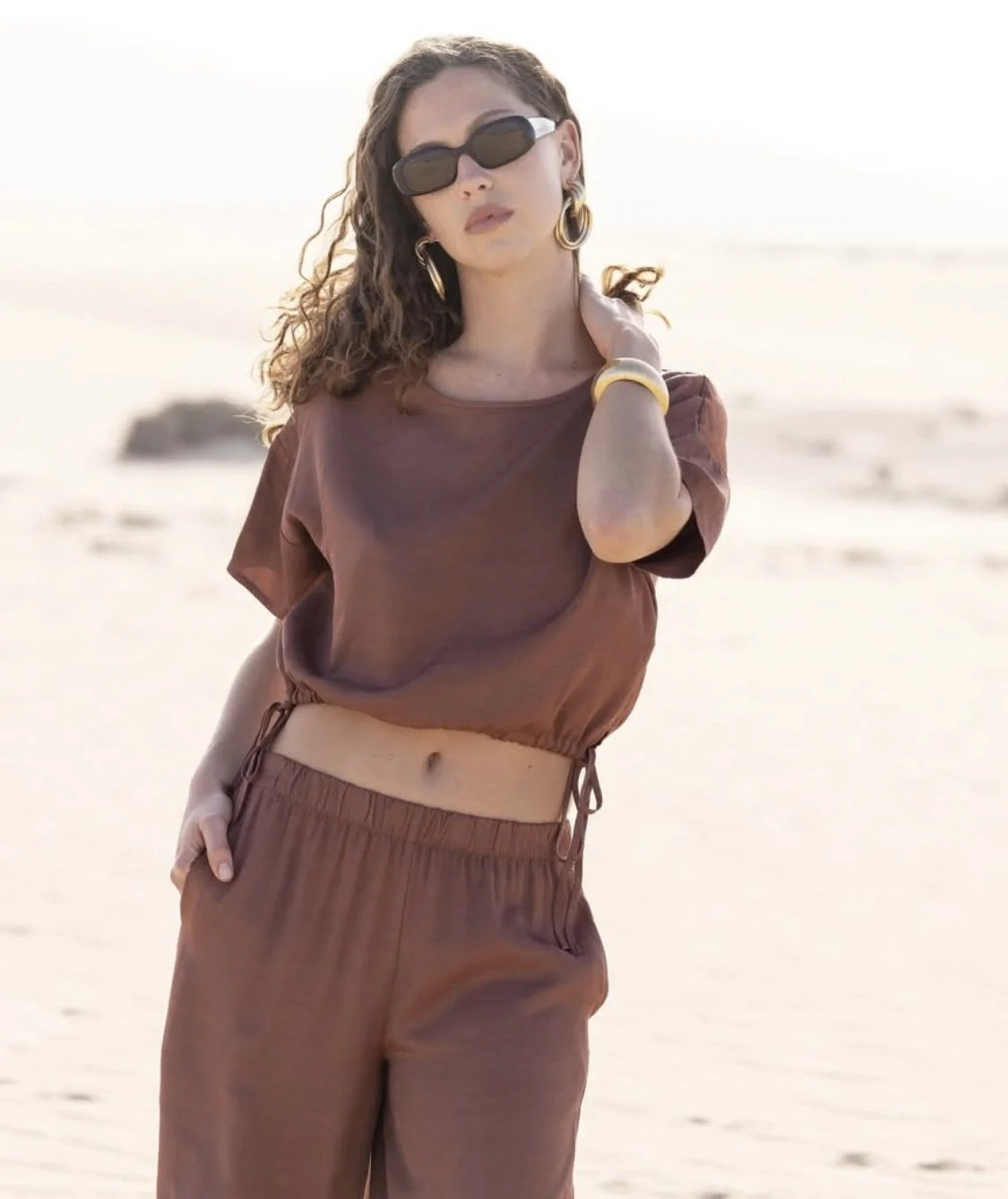Woman with curly hair wearing sunglasses, gold earrings, and jewelry, standing on a beach with sandy background, dressed in a brown loose crop top and matching brown pants.