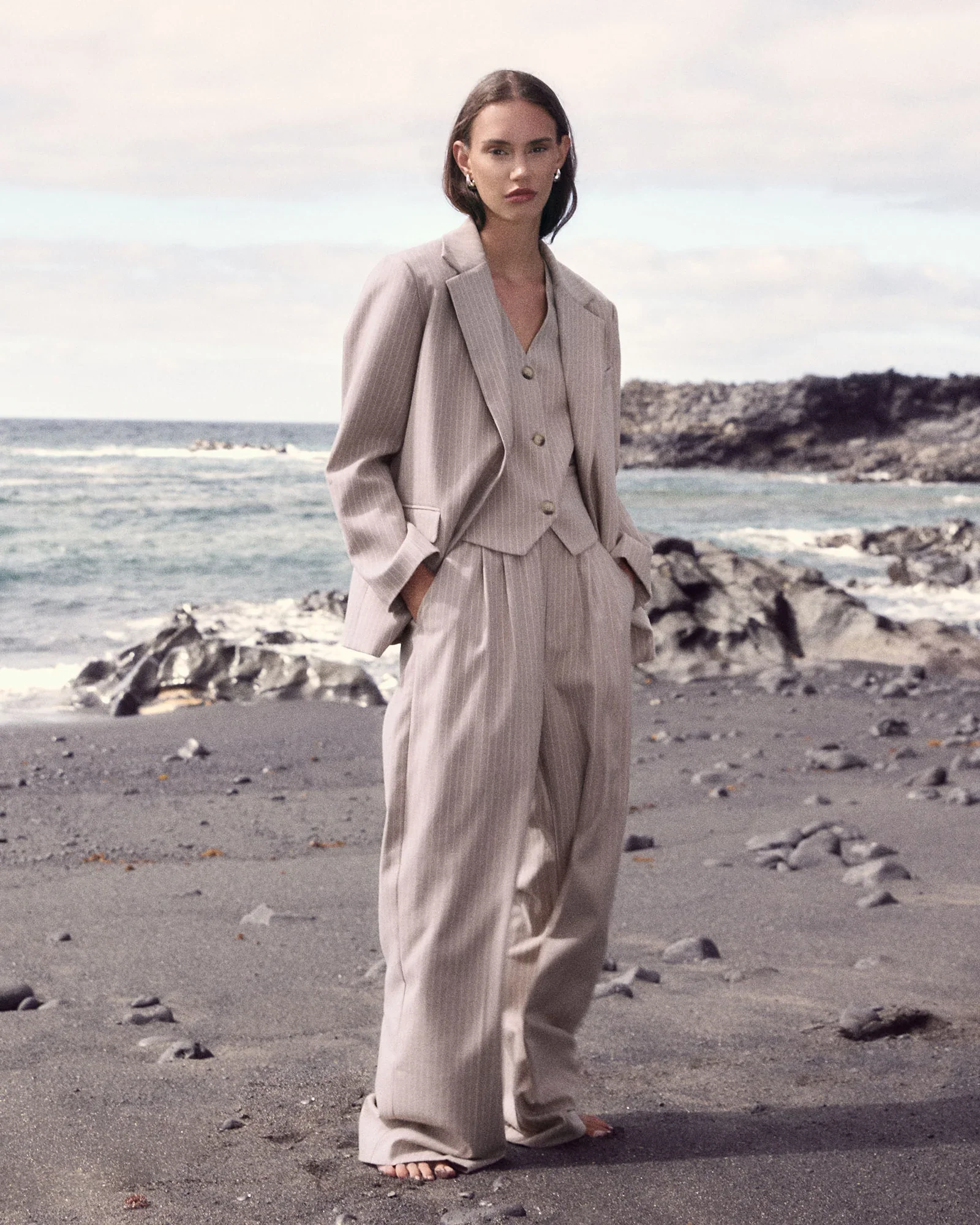 Woman in beige pinstripe suit standing on rocky beach with ocean in background