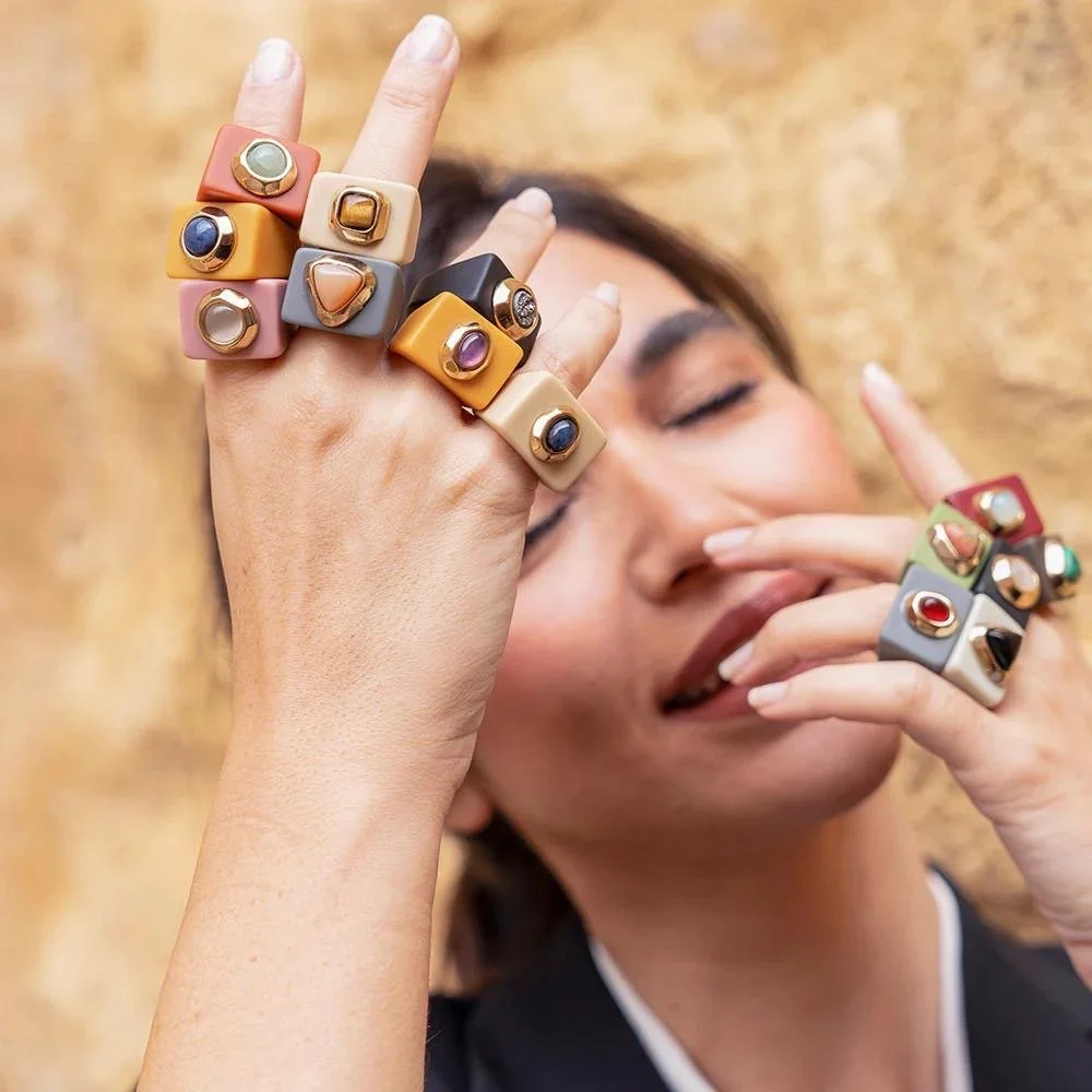 Woman showcasing colorful statement rings with various gemstones, holding her hand up near her face against a blurred beige background.
