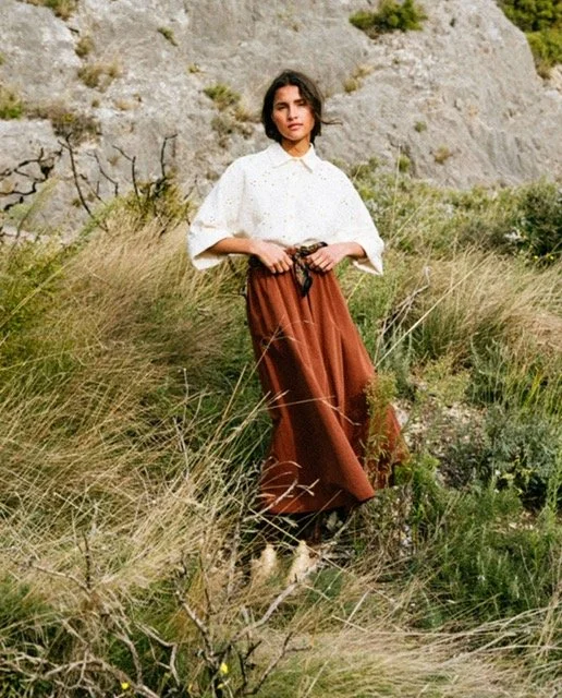 A woman standing outdoors among tall grass and shrubs with rocky hills in the background, wearing a white top and brown skirt.