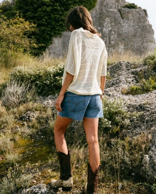 Woman standing outdoors with rocky terrain and greenery in the background, wearing a loose white knit sweater, denim shorts, and cowboy boots.