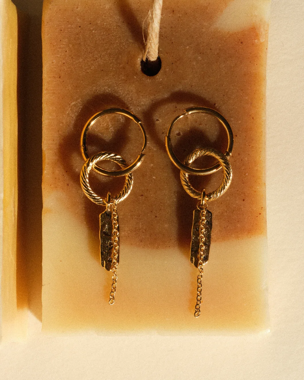 Pair of gold hoop earrings with dangling chain and textured metal charms, displayed on a pink foam pad.