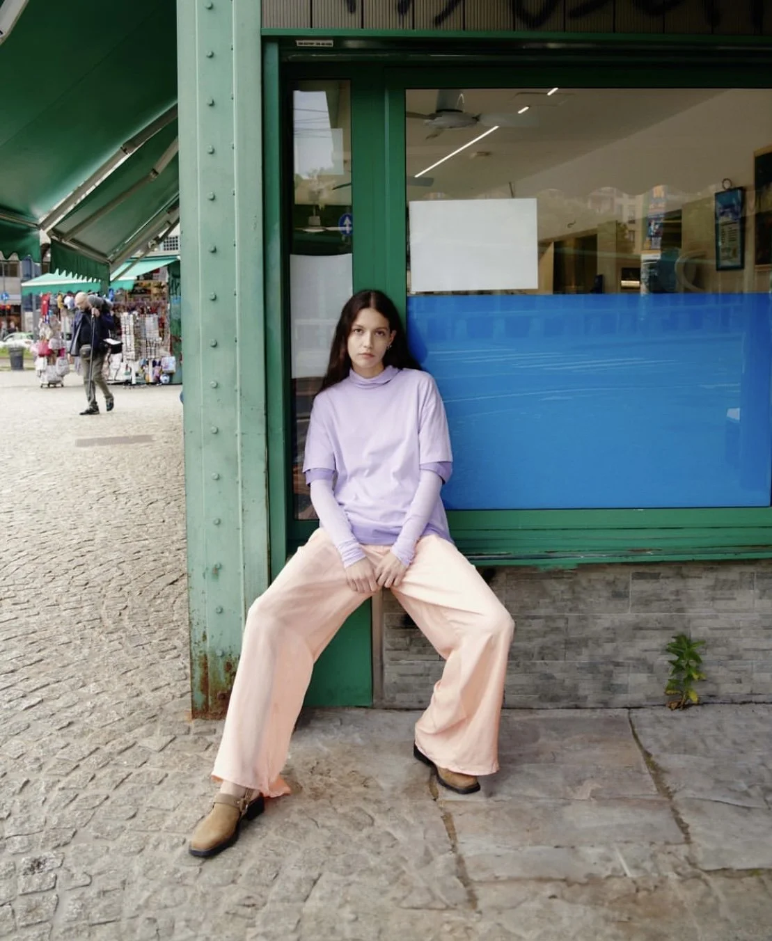 A young woman with long dark hair wearing a lavender long-sleeve shirt and loose pink pants sitting against a teal-colored shopfront with large windows in an outdoor market area.