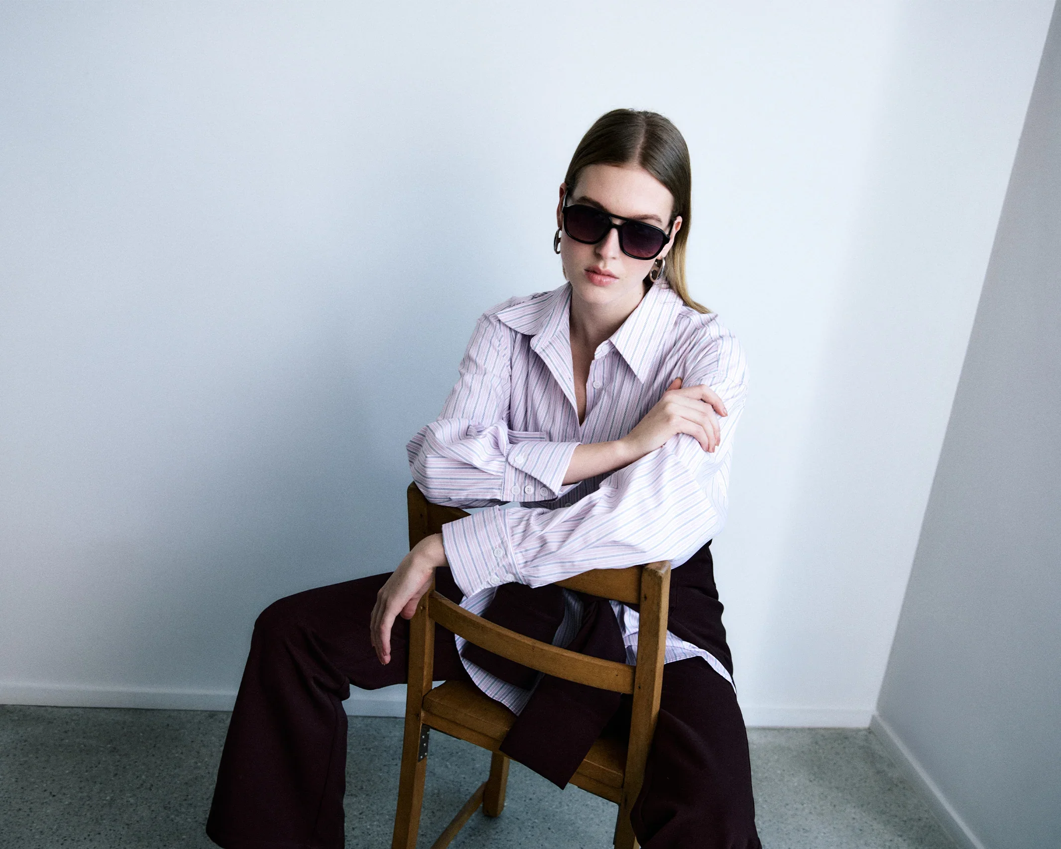 A woman with brown hair wearing sunglasses, a light striped shirt, and dark pants, sitting on a wooden chair against a plain white wall.