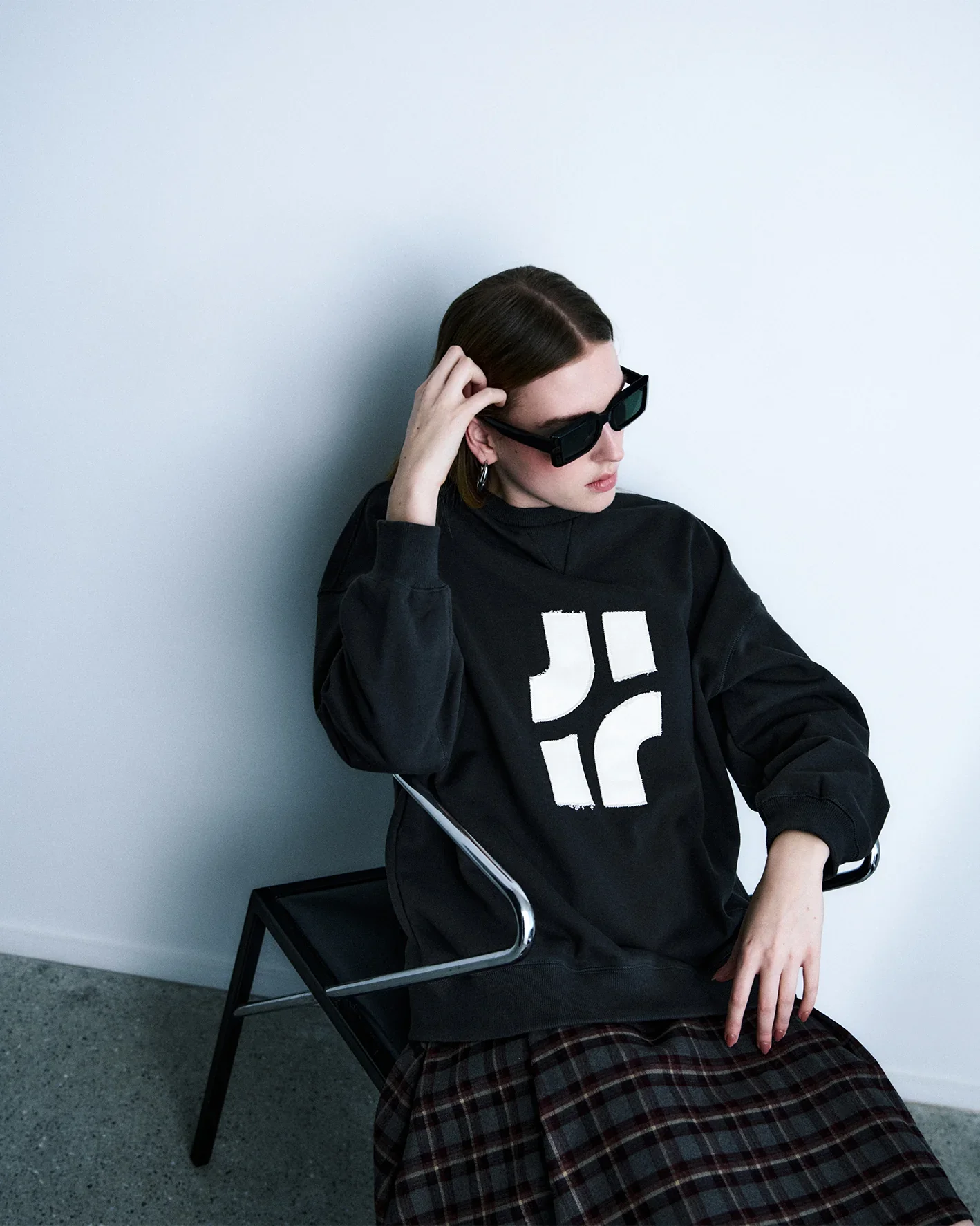 A woman with brown hair wearing black sunglasses, a black sweatshirt with white geometric design, and plaid skirt, sitting on a black chair against a plain white wall.