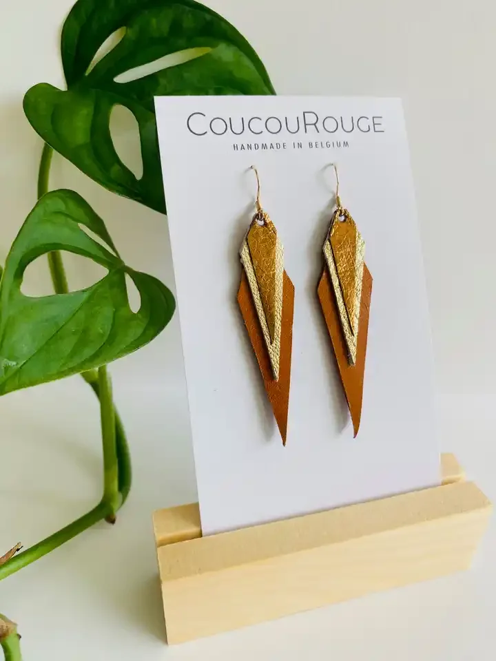 Gold and brown geometric dangle earrings displayed on a white card with the brand name 'CoucouRouge' and the text, 'Handmade in Belgium', resting in a wooden stand. A green plant with large leaves is beside the display.