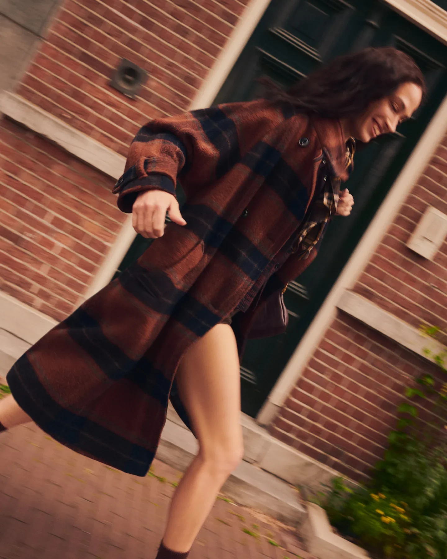 Woman in a plaid coat running, with the coat open revealing her legs, near a brick wall and green door.