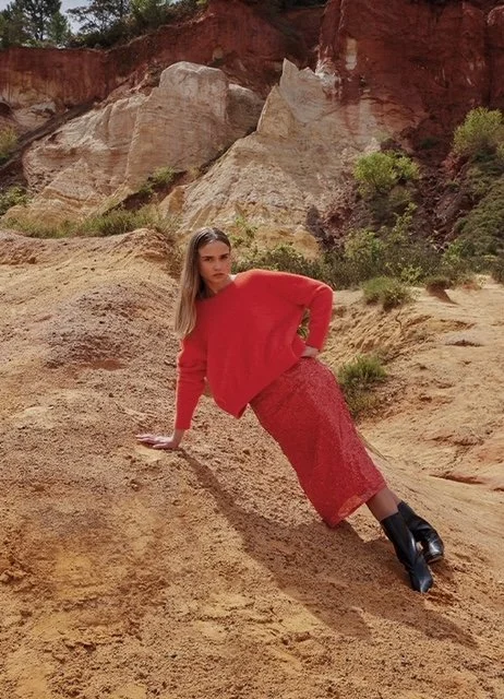 A woman in a red oversized sweater and red skirt with black boots, leaning on a sandy slope in a desert landscape with reddish rock formations and sparse green bushes.