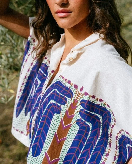 A woman wearing a white shirt with colorful patterns of animals and plants, standing outdoors in a natural setting.