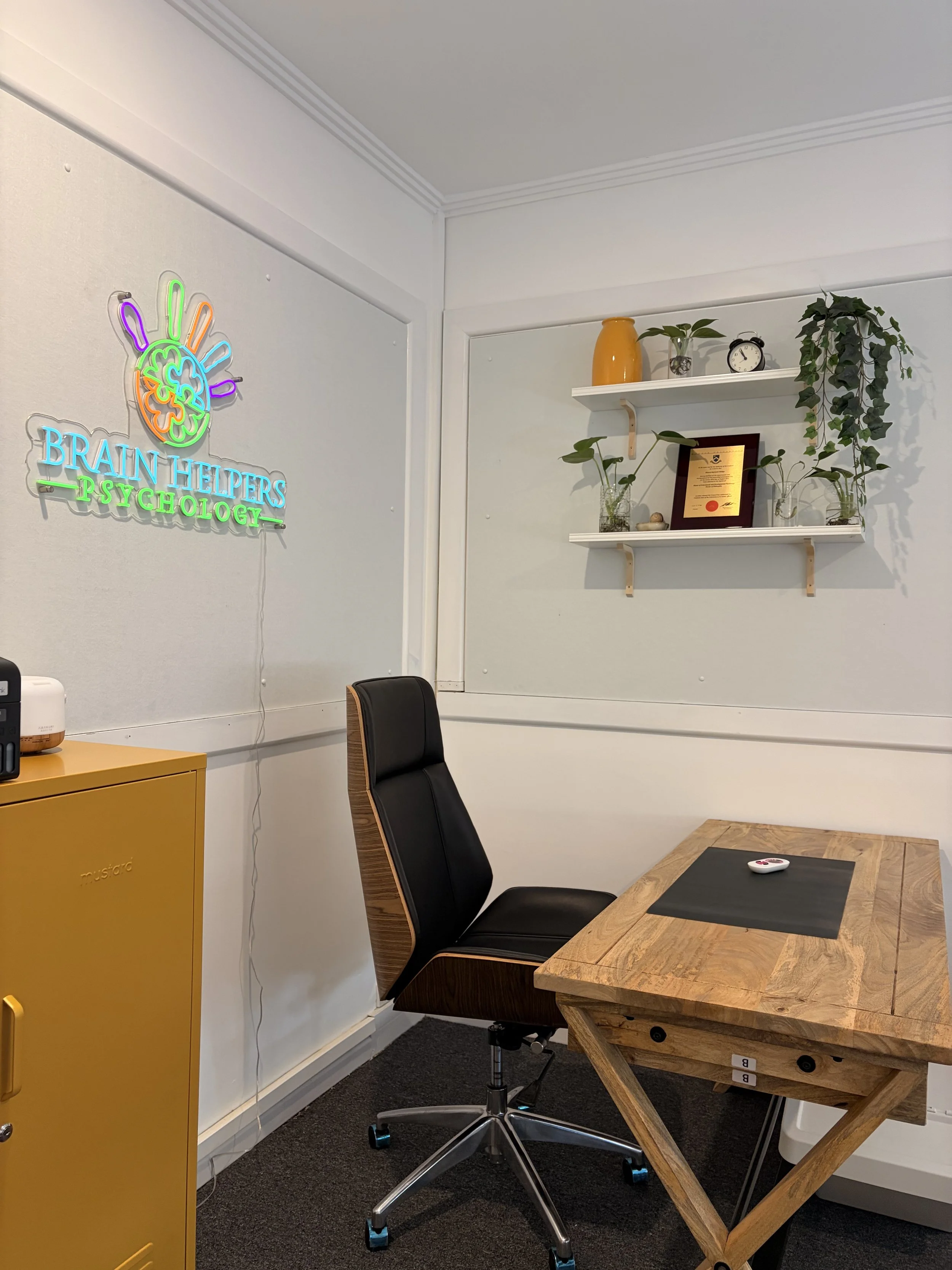 An office with a black rolling chair, a wooden desk with a black pad and white device, a yellow cabinet, white walls with a neon sign reading 'BRAIN HELPERS PSYCHOLOGY,' and two white shelves decorated with plants, a framed certificate, a yellow vase