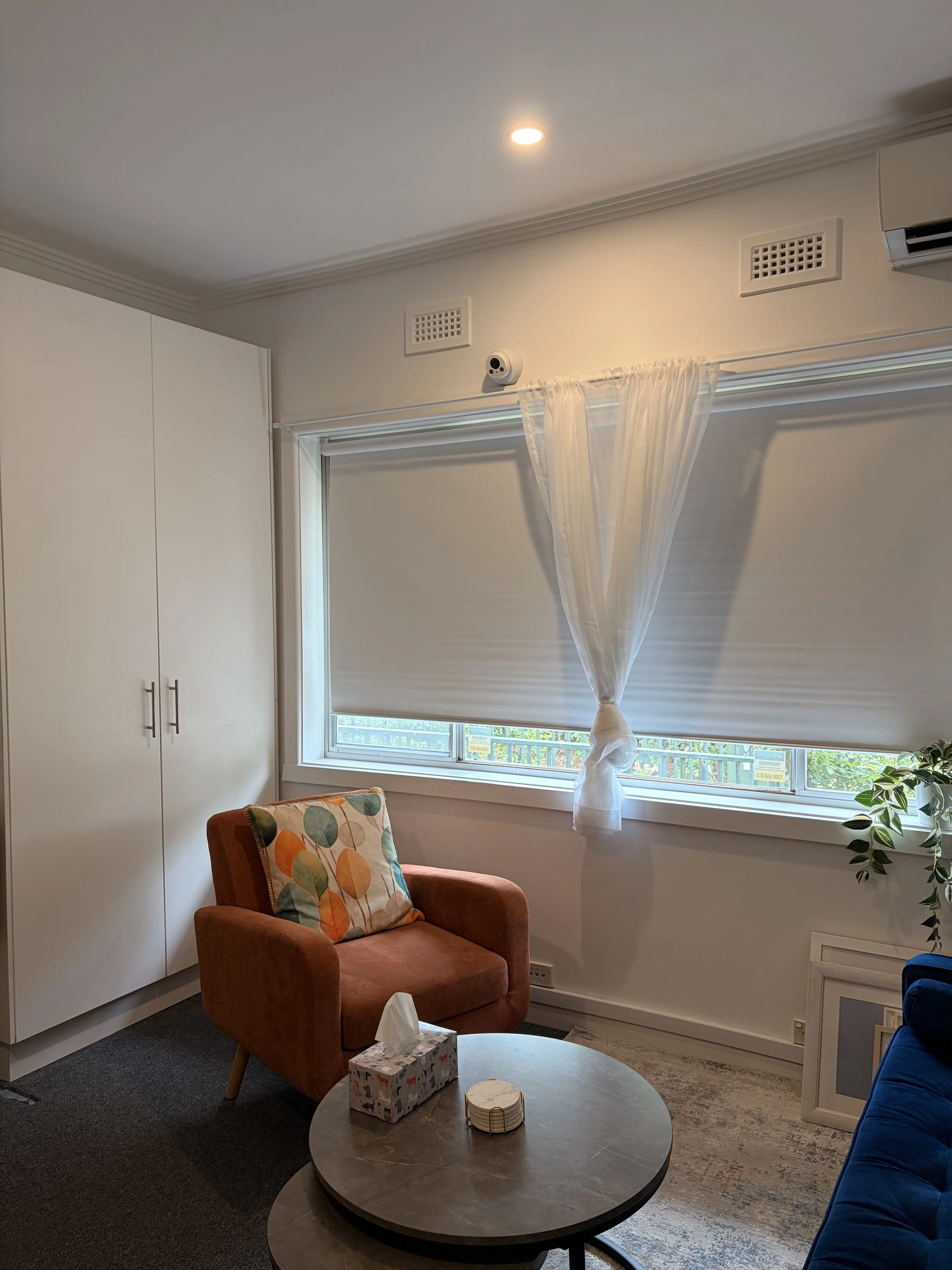 Living room with a rust-colored armchair, a small round coffee table with tissue box and coasters, white walls, window with white roller shade and sheer curtain, potted plant, corner of a blue sofa, storage cabinet, ceiling light, and wall vents.