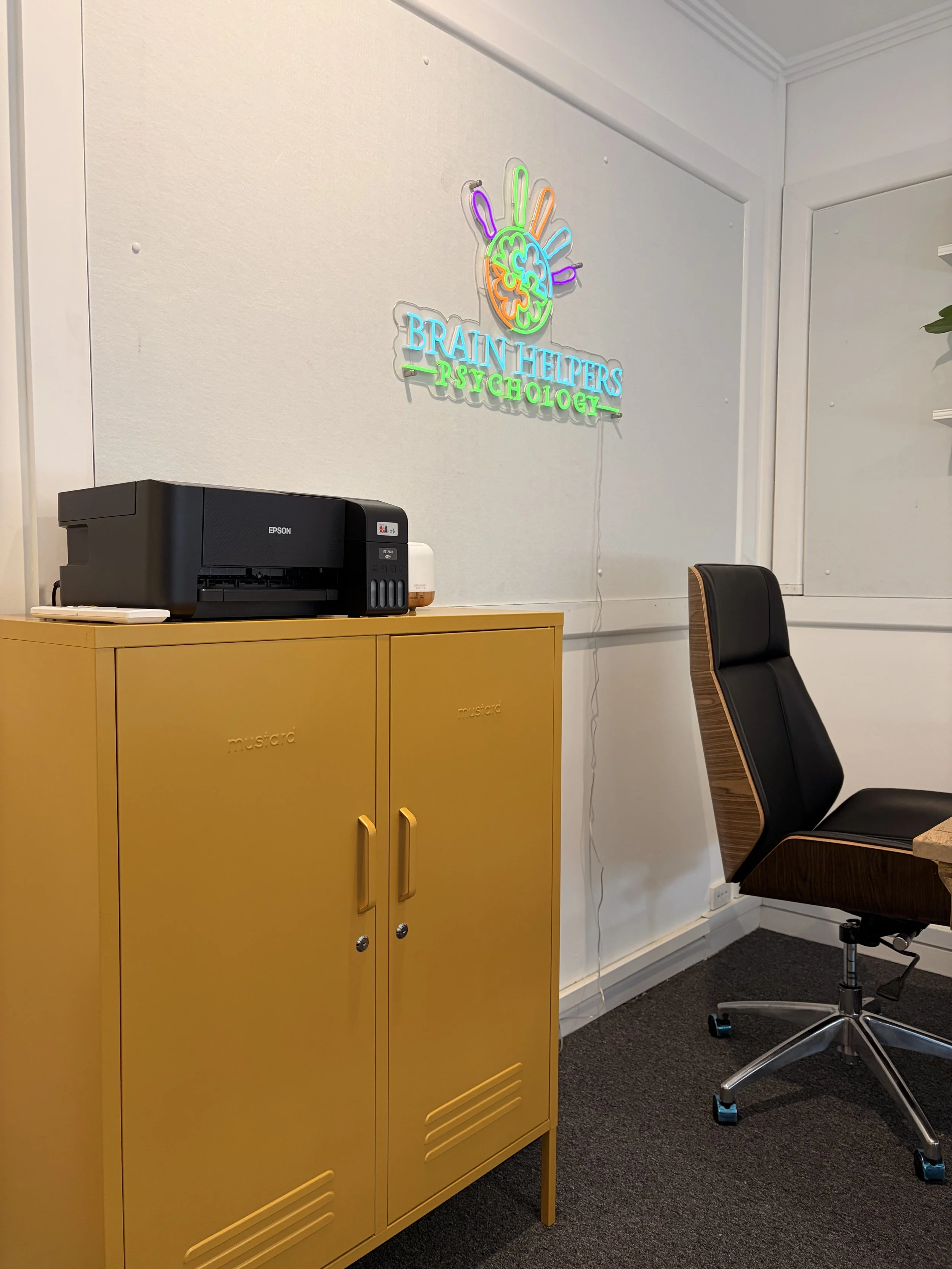 Office space with a black Epson printer on a yellow cabinet, a black office chair, and a neon sign on the wall that reads 'Brain Helpers Psychology' with a colorful brain and hand design.