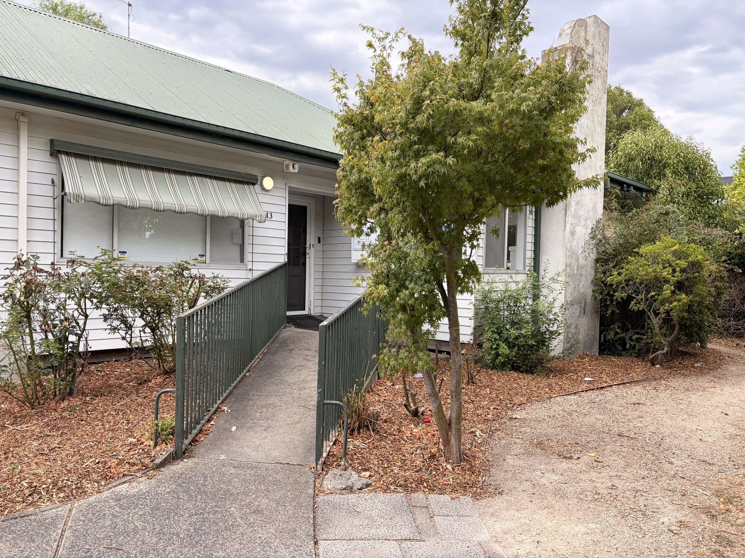 Exterior of a white house with a small ramp leading to the front door, surrounded by trees and bushes. The house has a green roof and a striped awning over a window.