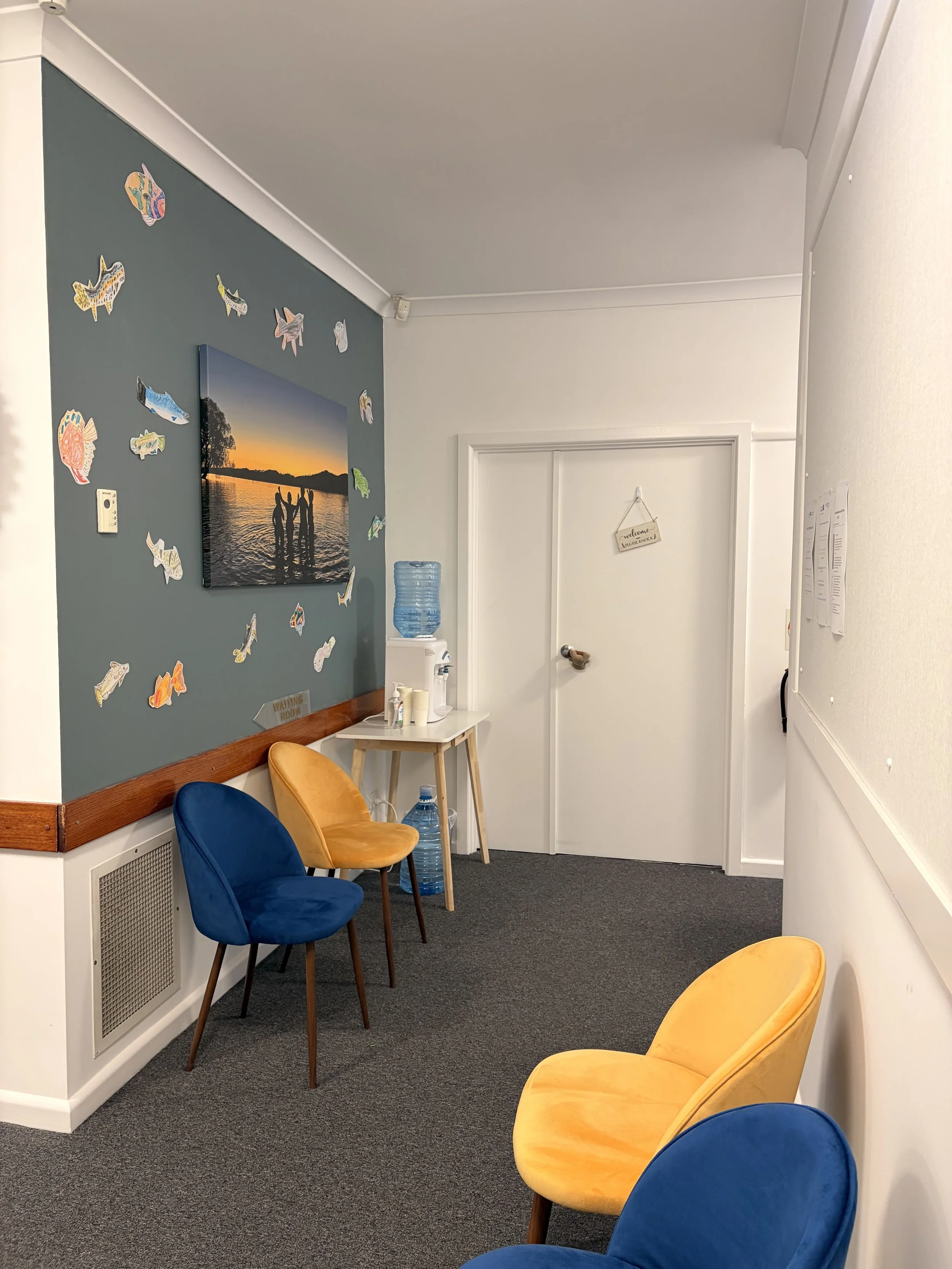 A waiting room with blue and yellow chairs, a water dispenser, a large water bottle on the floor, and a wall decorated with fish cutouts and a sunset photo.