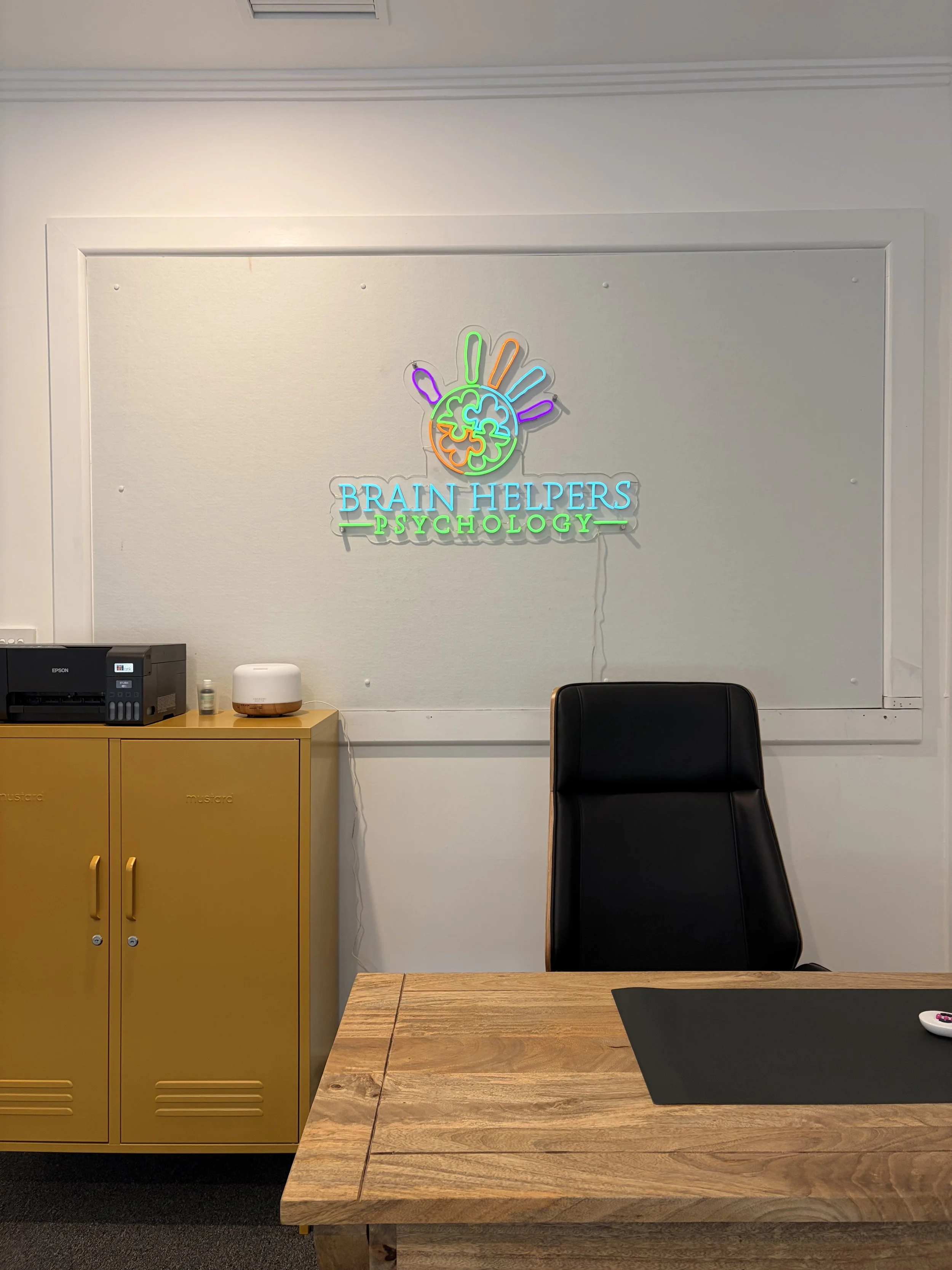 Neon sign on a white wall that reads 'Brain Helpers Psychology' with a colorful brain and hand logo. Office desk and black leather office chair in the foreground.