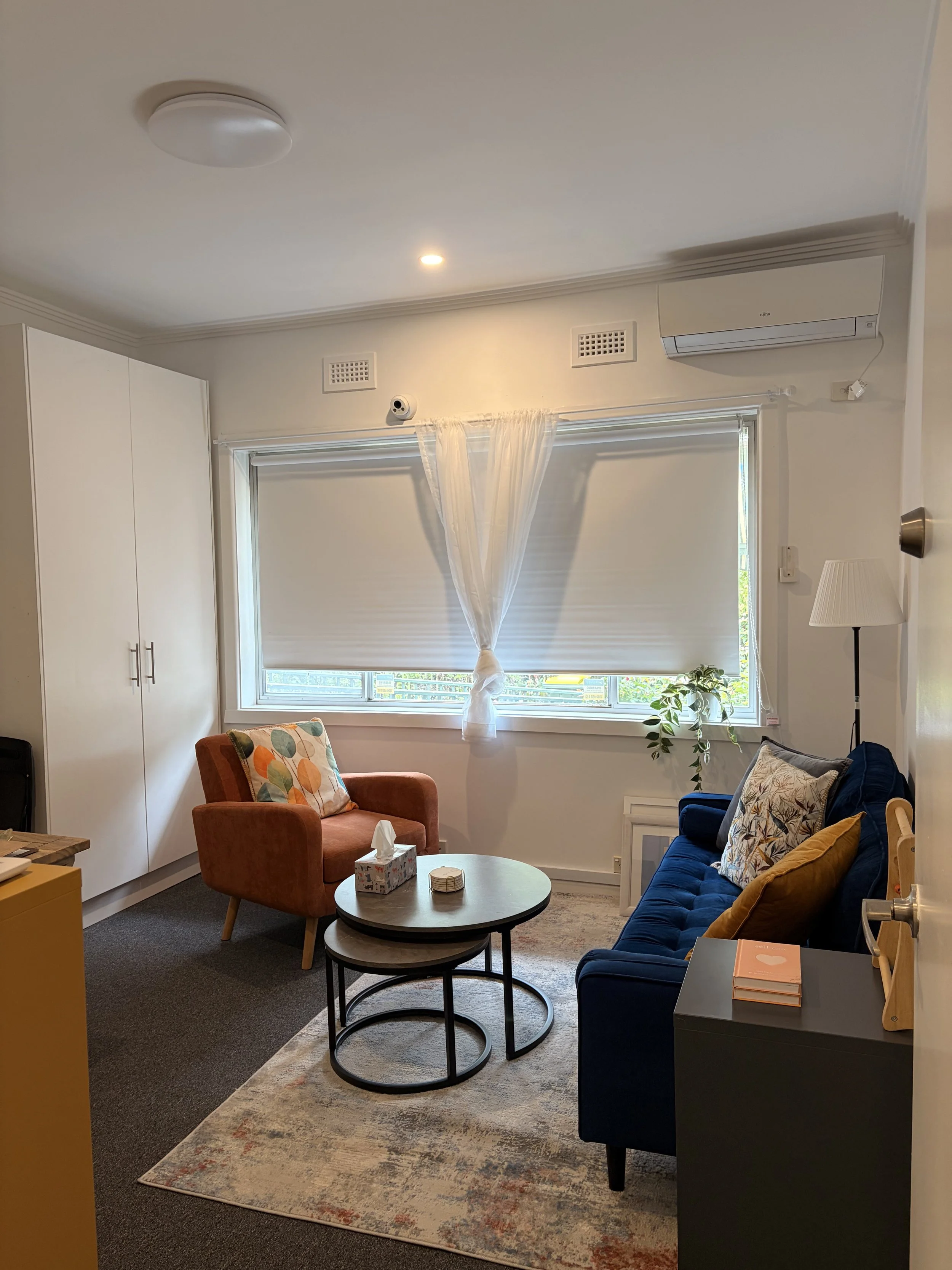 Living room with a navy blue sofa, a coral armchair, a round nested coffee table, a white curtain, and a window with blinds. There's a potted plant on the windowsill, a standing lamp, a white wall-mounted air conditioner, and a carpeted floor.