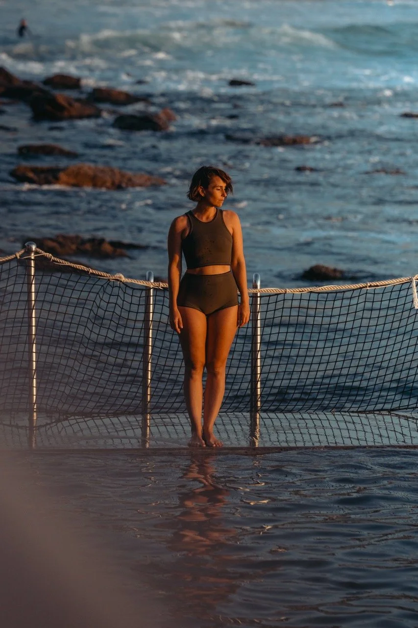 A woman standing by a railing at the edge of a body of water, possibly at the beach during sunset, wearing a black swimwear