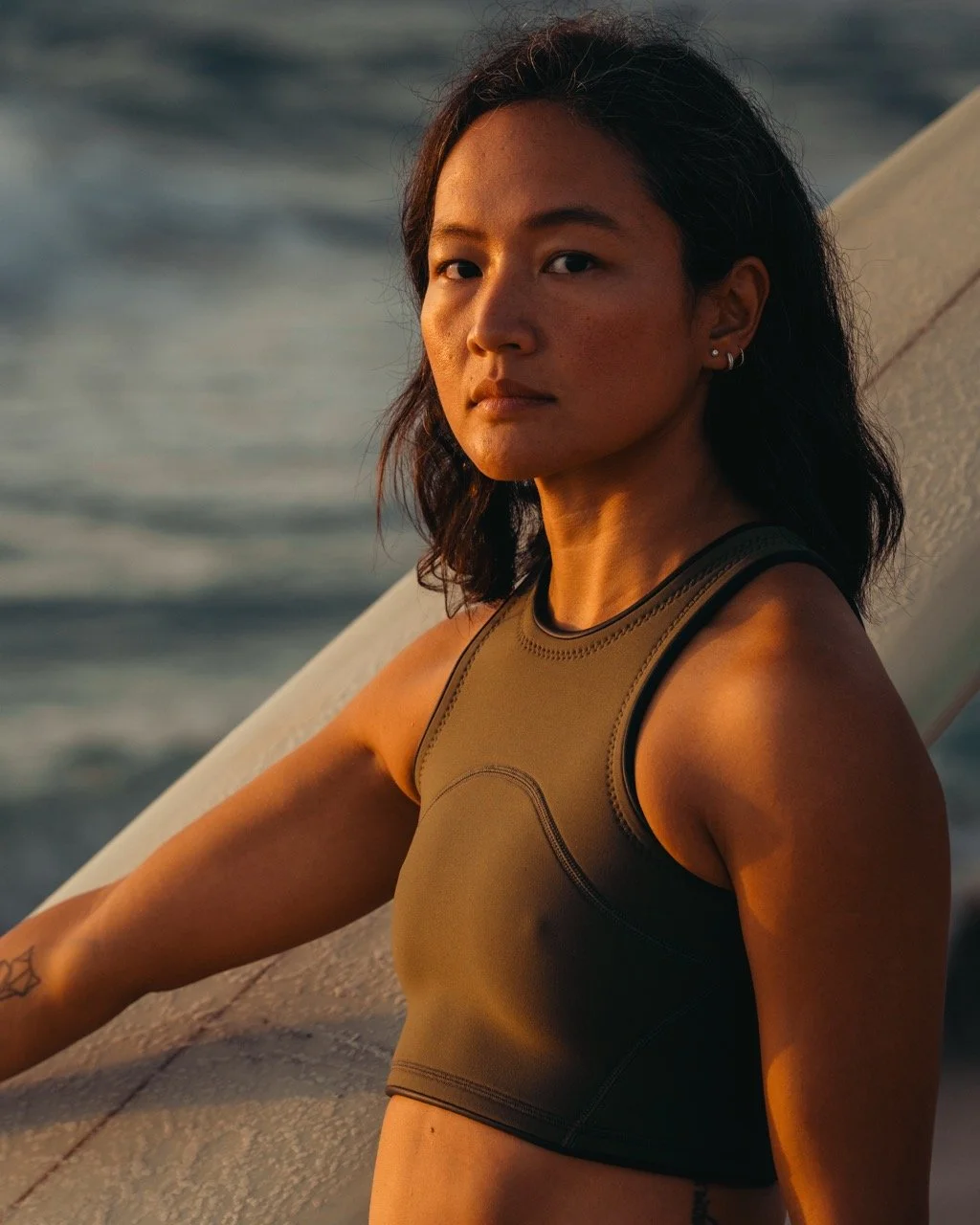 A young woman with dark hair and earrings looking at the camera outdoors during sunset, near water, wearing a sleeveless athletic top.
