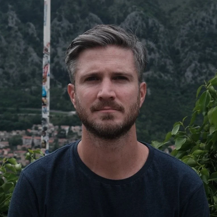 A man with short brown hair and a beard, wearing a black shirt, standing outdoors with green foliage, mountains in the background, and a pole with stickers.
