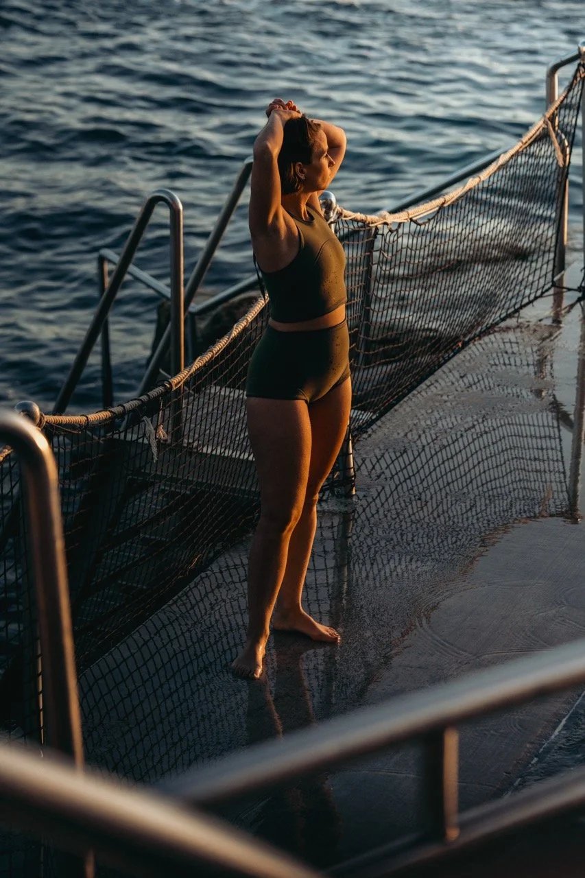 A woman in a black sports bra and shorts standing on the deck of a boat, looking out at the water with her hands behind her head during sunset.