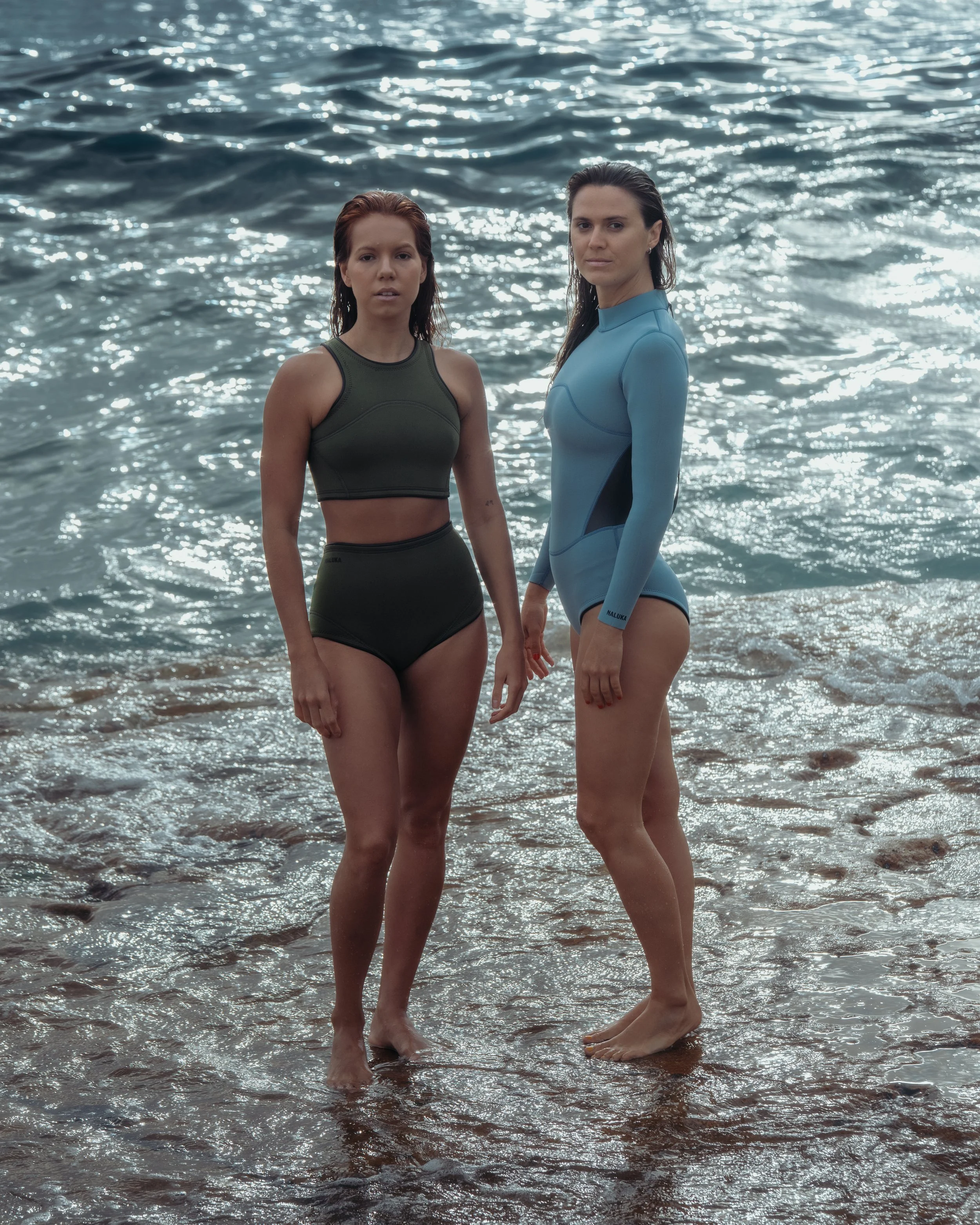 Two women in wetsuits standing on a beach with water in the background.