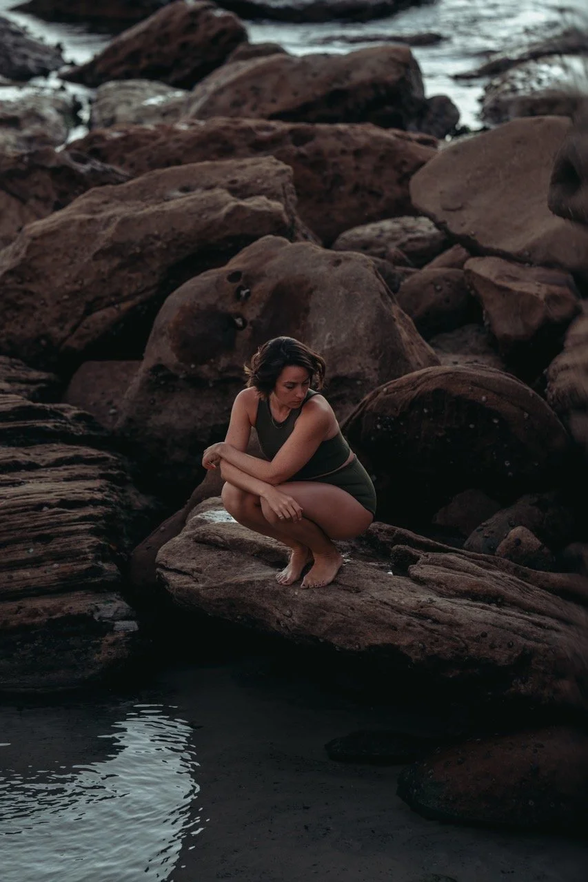 A woman with dark hair squats on a large rock by a body of water, surrounded by dark brown rocks.