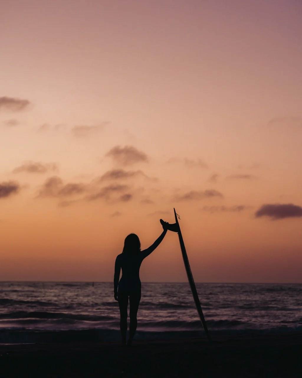 Silhouette of a woman standing on the beach at sunset, holding a surfboard upright.