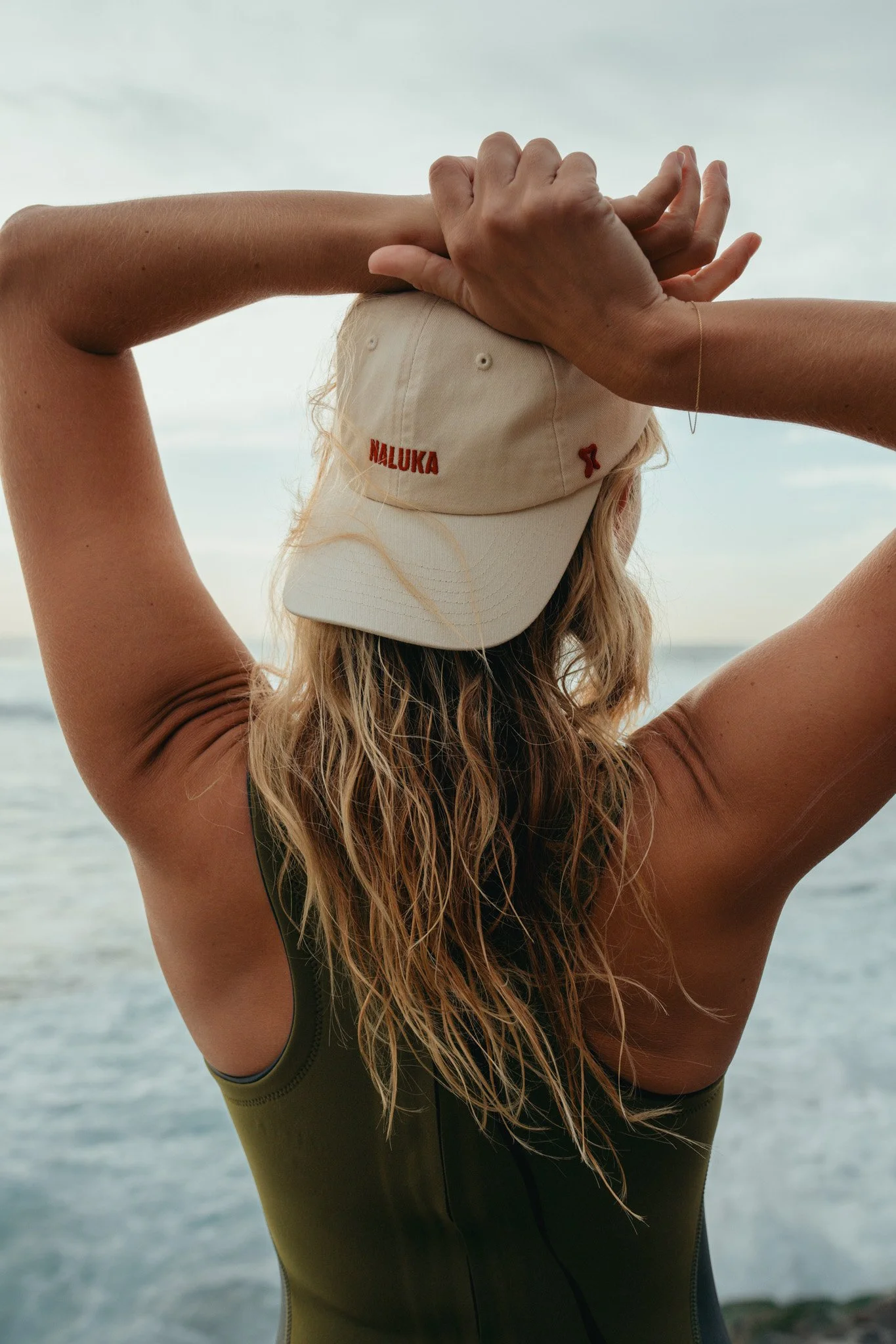 A woman with wavy blonde hair is standing outdoors near water, wearing a beige cap with red embroidery and a dark green sleeveless top, with her arms raised and hands clasped behind her head.